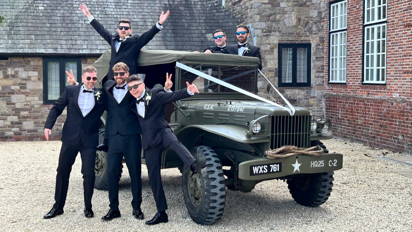 Groomsmen cheering and posing for photos around a Khaki Green Military Dodge Car. all groomsmen are wearing matching dark blue suits