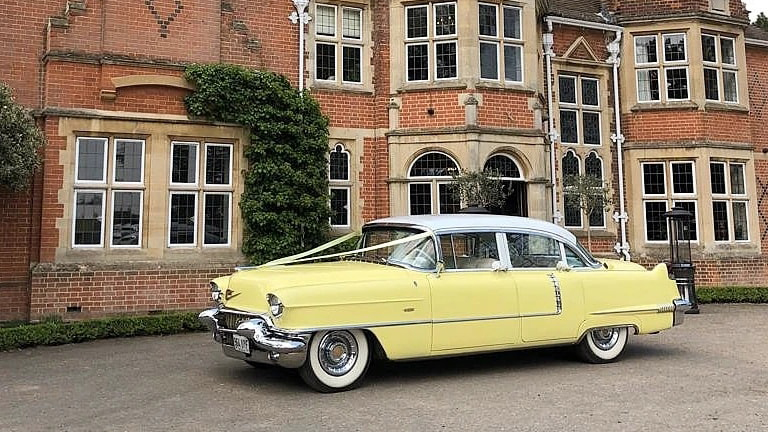 Yellow Classic Cadillac with white roof and decorated with White ribbons parked in front of wedding venue