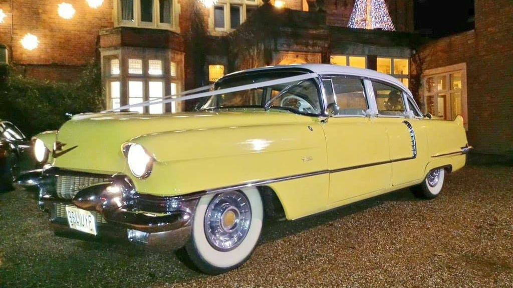 Classic Yellow Cadillac with white ribbons parked in front of wedding venue in the evening with headlights on