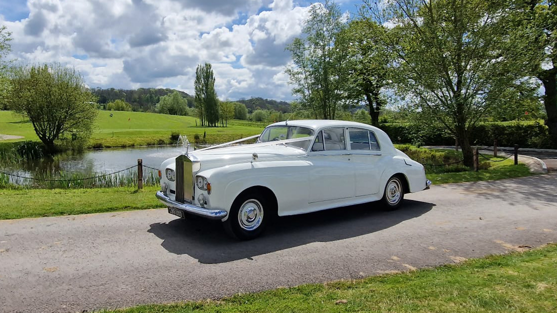 Traditional Classic Rolls-Royce dressed with white ribbons entering wedding venue in Wales with lac in the background