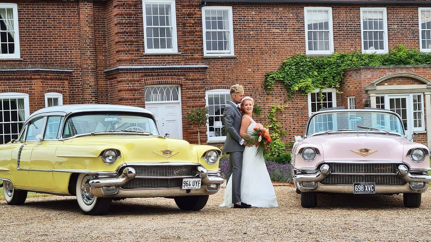 Bride and Groom posing for photos in the middle of two classic Cadillacs. The right car is pink and the left one is yellow