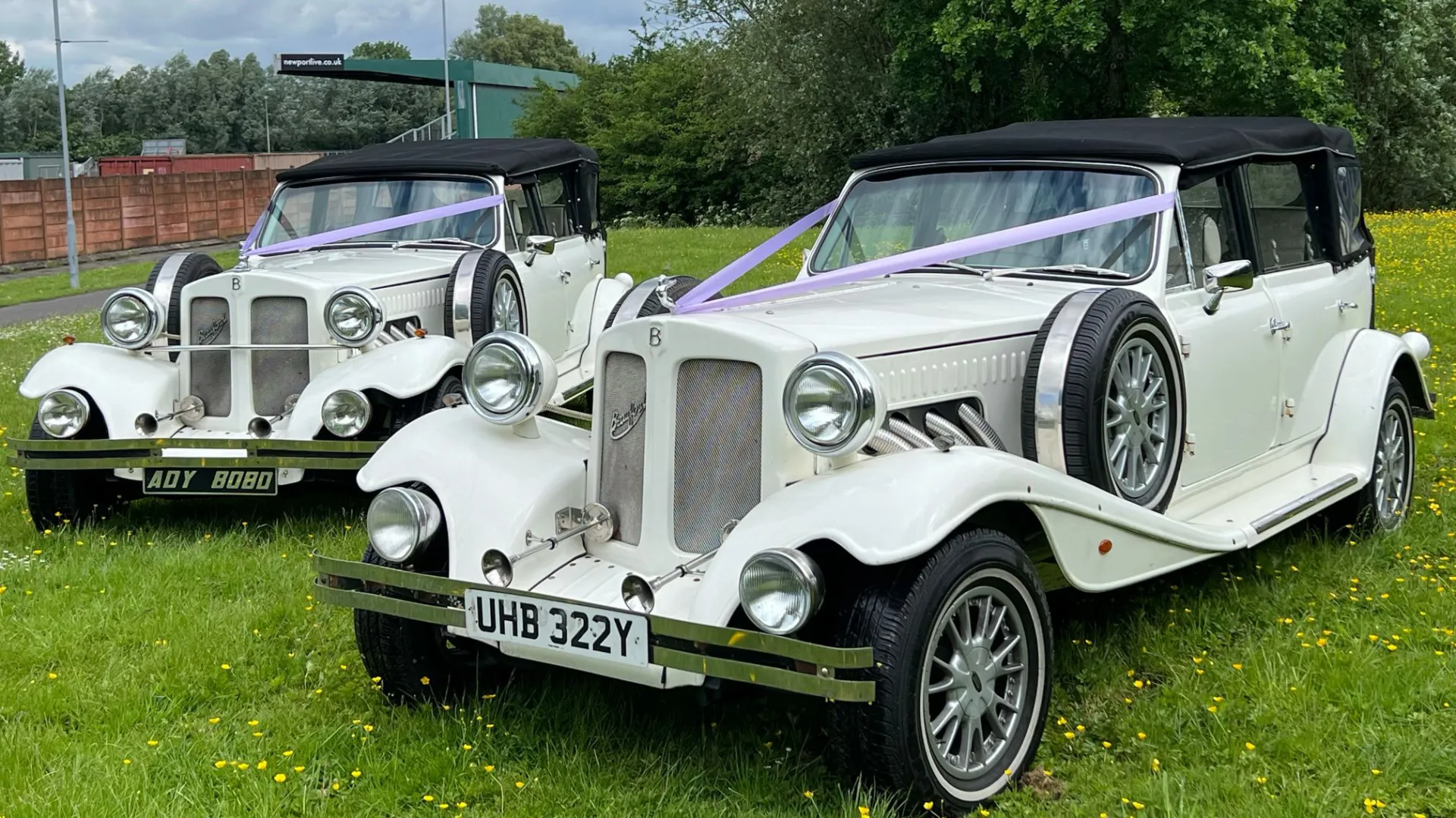 Two matching pair of Beauford Convertible parked in a green field and decorated with wedding ribbons