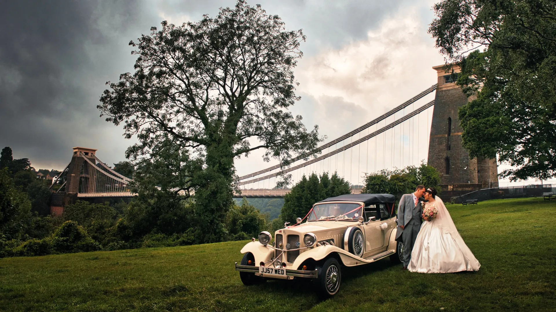 Vintage Beauford Convertible in Ivory decorated with wedding ribbons with Bride and Groom standing by the vehicle. Clifton Suspension Bridge in Bristol can be seen in the background