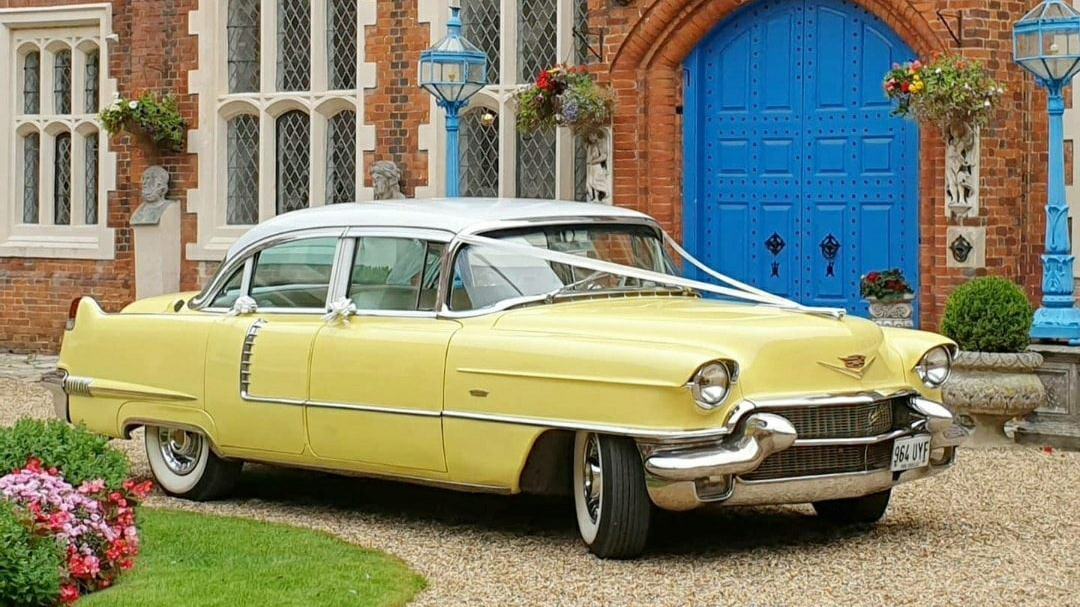 Yellow Cadillac with white ribbons parked in frotn of a wedding venue with large blue door.