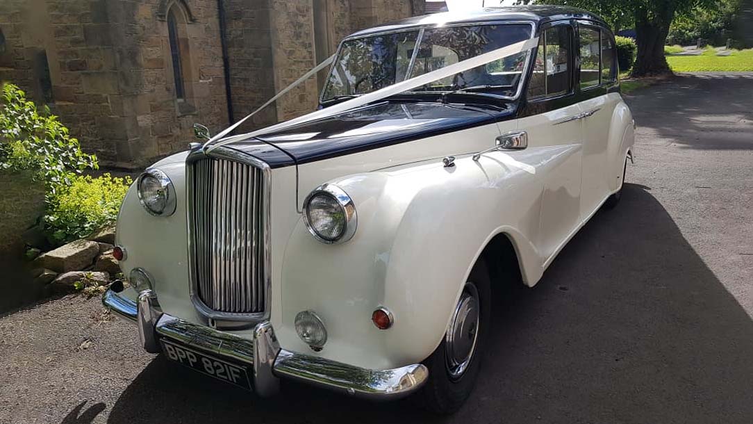 Ivory Austin Princess with Black Roof and bonnet decorated with White ribbons parked in front of a church