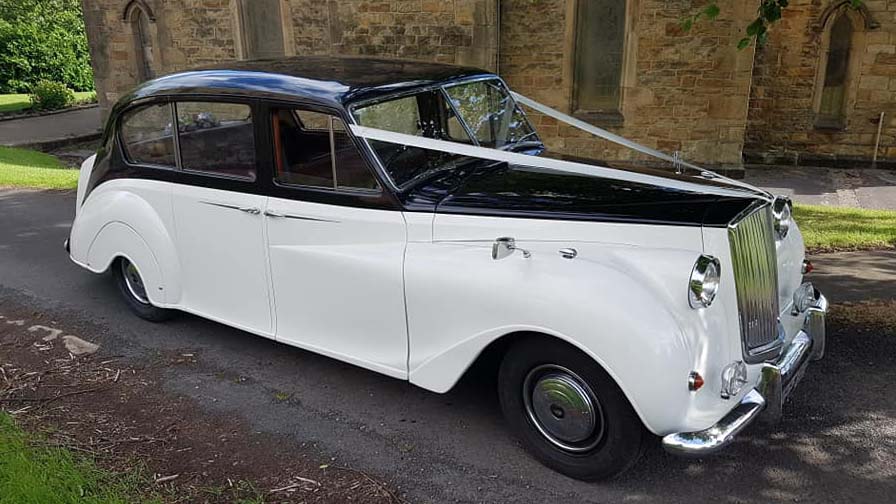 Right side vioew of Classic Austin Princess in Ivory with Black Roof and Bonnet decorated with White Ribbons parked n front of a Church