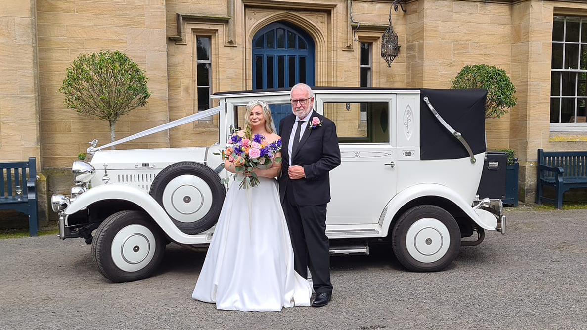 White Imperial Convertible parked in front of wedding venue with Bride and Groom posing for photos in front of the vehicle.