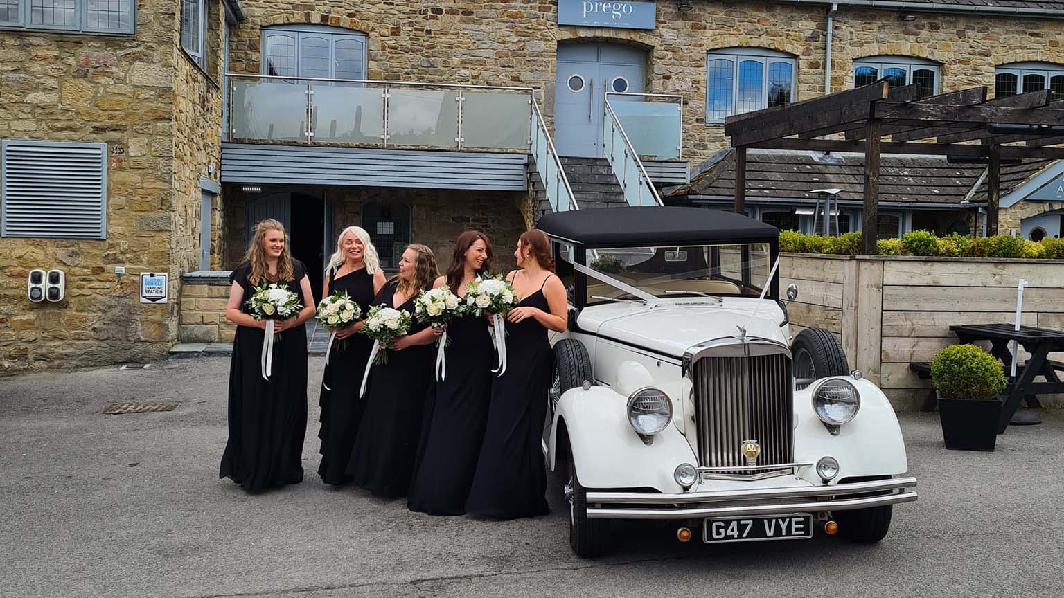 5 Bridesmaids wearing black dresses and holding matching flowers in their hands all lined up in front of Regent Convertible wedding car