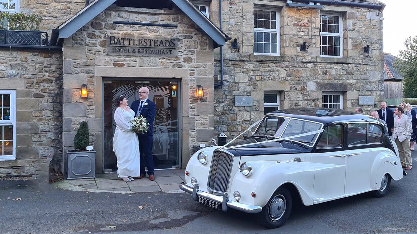 Classic Black and Ivory Austin Princess decorated with White ribbons accross its black bonnet  parked in front of wedding. Bride and Groom are holding hands in front of the venue's main Entrance