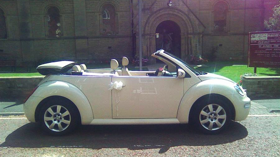 side view of VW Beetle with convertible roof down and white bows on the door handle parked in front of a church