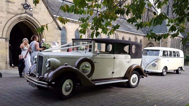 Vintage Regent Convertible folowed by a VW campervan dressed with matching white ribbons parked in front of Church. Bridal party can be seen in the background.