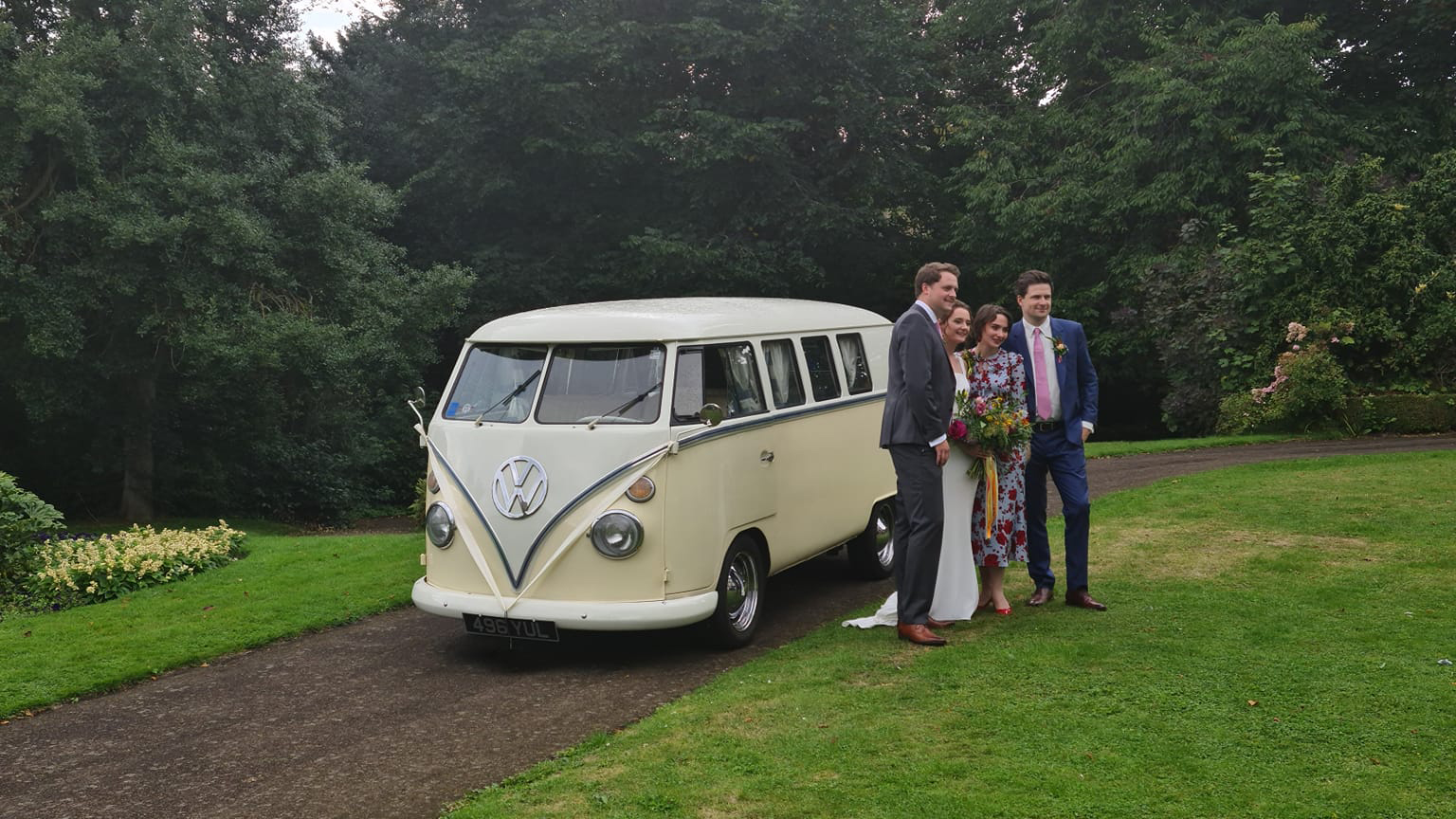 Classic VW Campervan parked on drive surrounded by green grass and trees with Bride , groom and parents standing by the vehicle