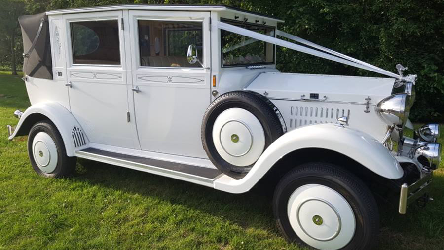 Right view of vintage style imperial with black soft top roof closed and spare wheel mounted on the side of the vehicle. Vehicle is decorated with white ribbons accross it bonnet