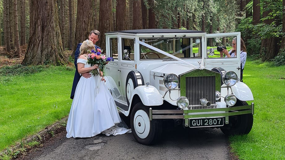 White vintage sytle imperial wedding car decorated with white ribbons parked accross  a path with bride and groom standing by the vehicle kissing