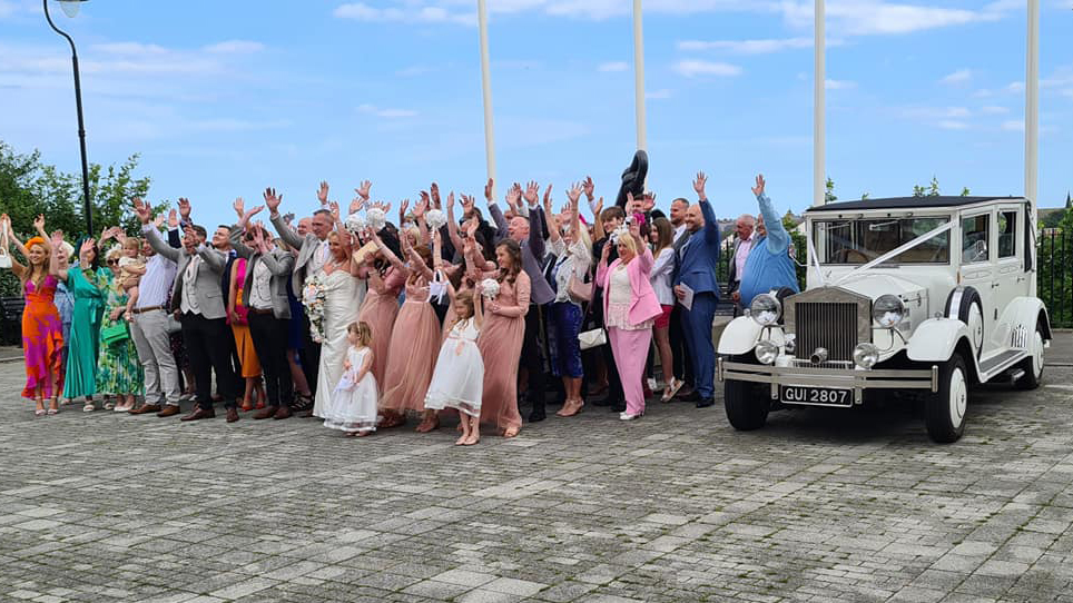 Full wedding party cheering with their hands up next to a vintage Imperial wedding car decorated with ivory ribbons