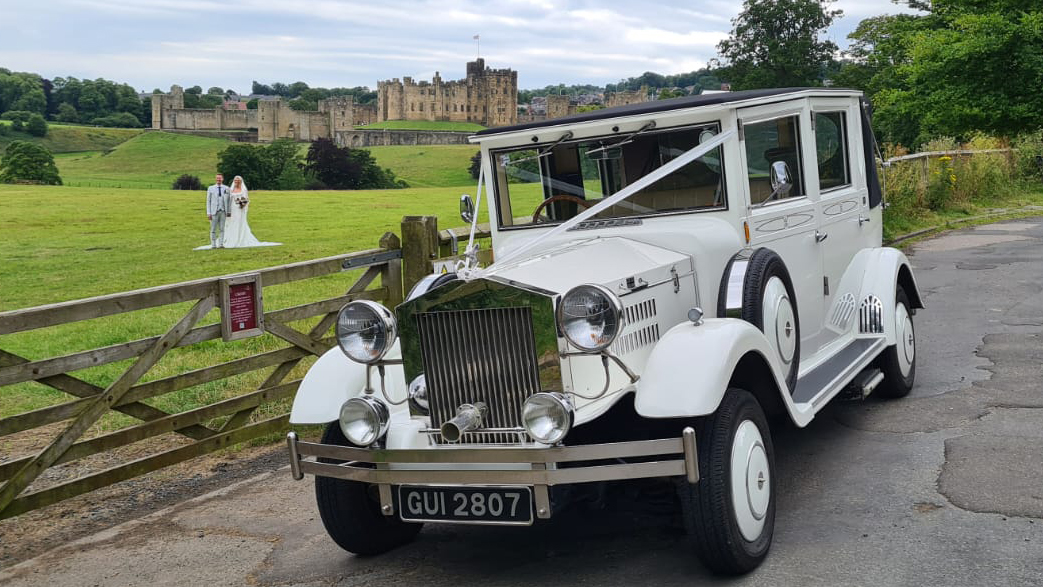 White Imperial wedding car parked on the side of the road. Bride and groom can be seen in the far background having their photos taken