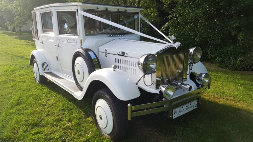 Right front view of White Imperial Convertible in a field with green trees in the background. Vehicle is decorated with White ribbons, large chrome radiator.