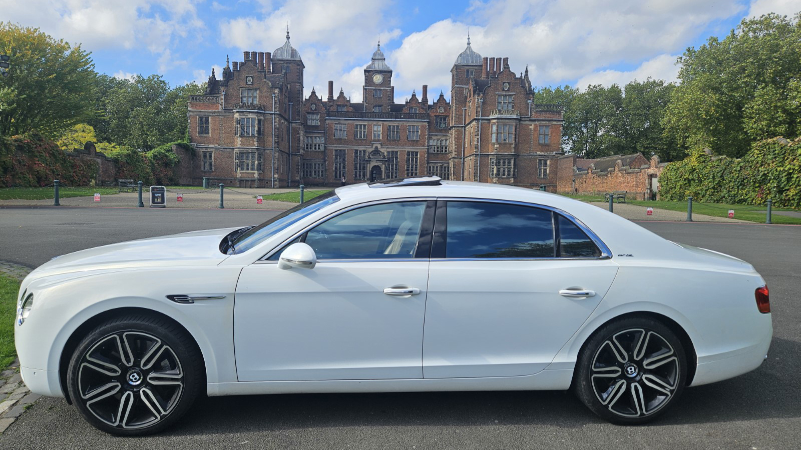 Left side view of Modern White Bentley with large Black and Silver Alloy wheels, sunroof open. Wedding venue in the far background