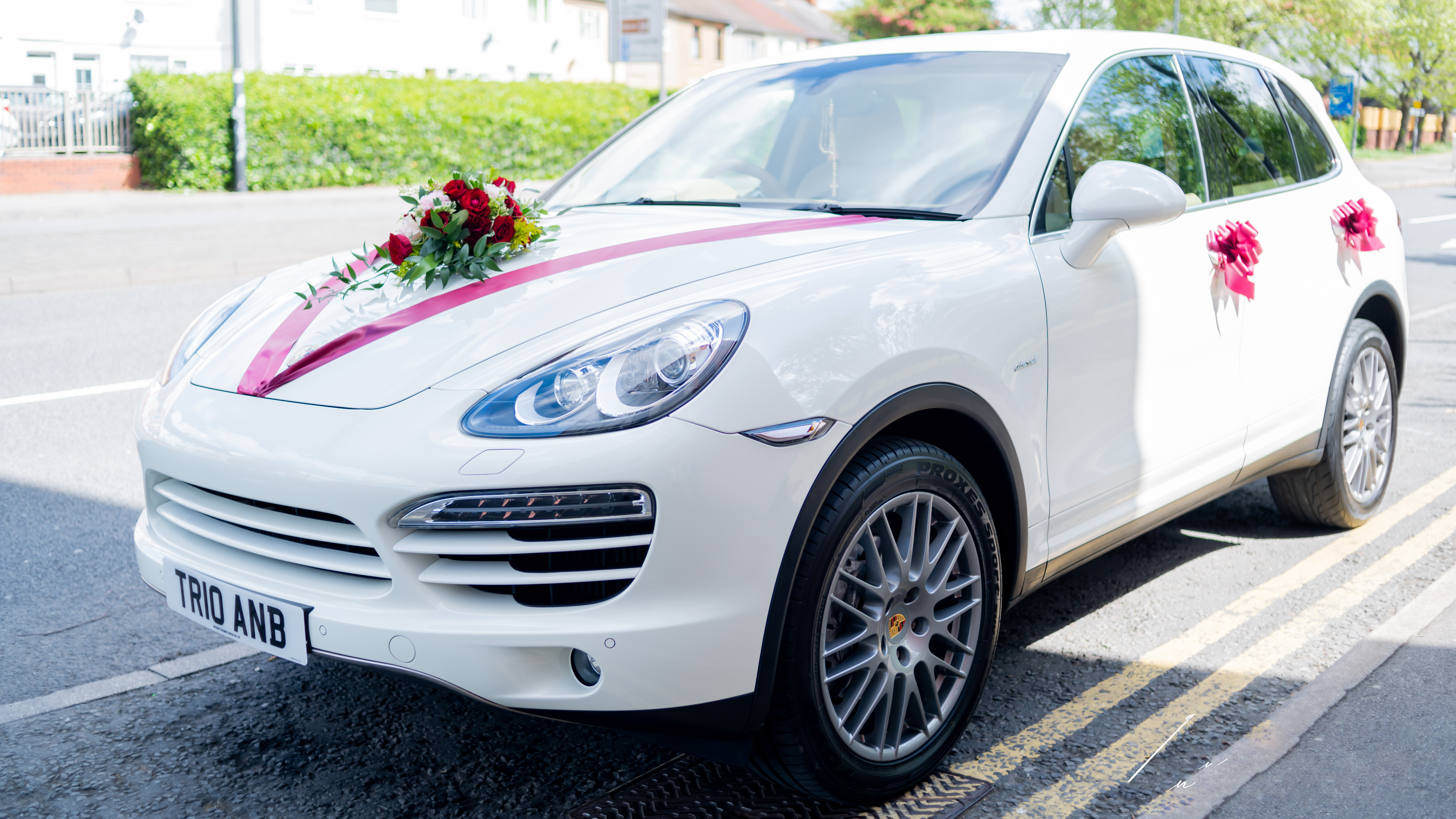 Modern white Porsche Cayenne parked on the side of the street with Burgundy ribbons accross front bonnet, matching burgundy bows on door handles and posy of flowers on the bonnet.