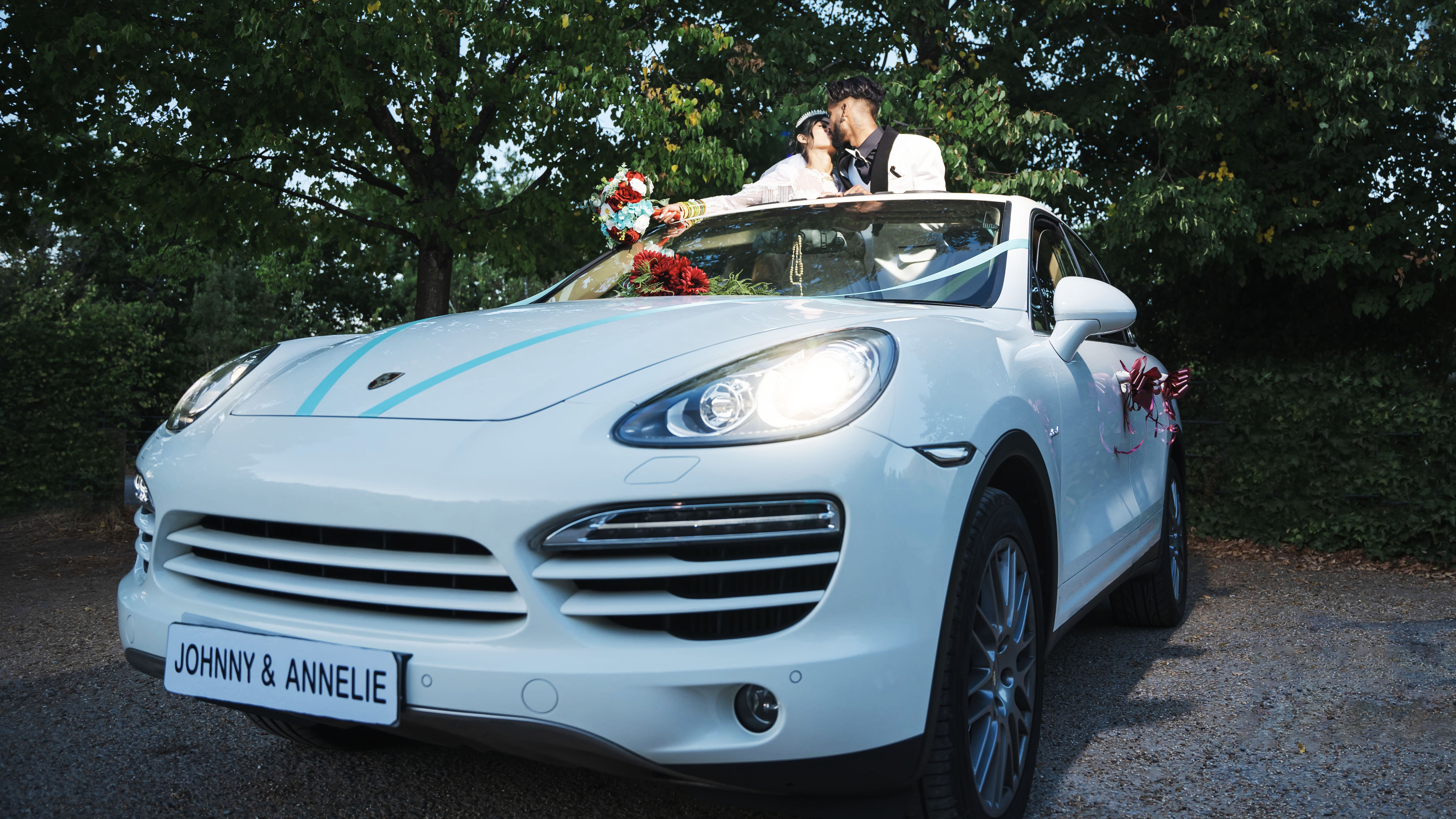 White Porsche Cayenne with light blue ribbons, headlights on. Bride and Groom are standing in the vehicle through the sunroof kissing.