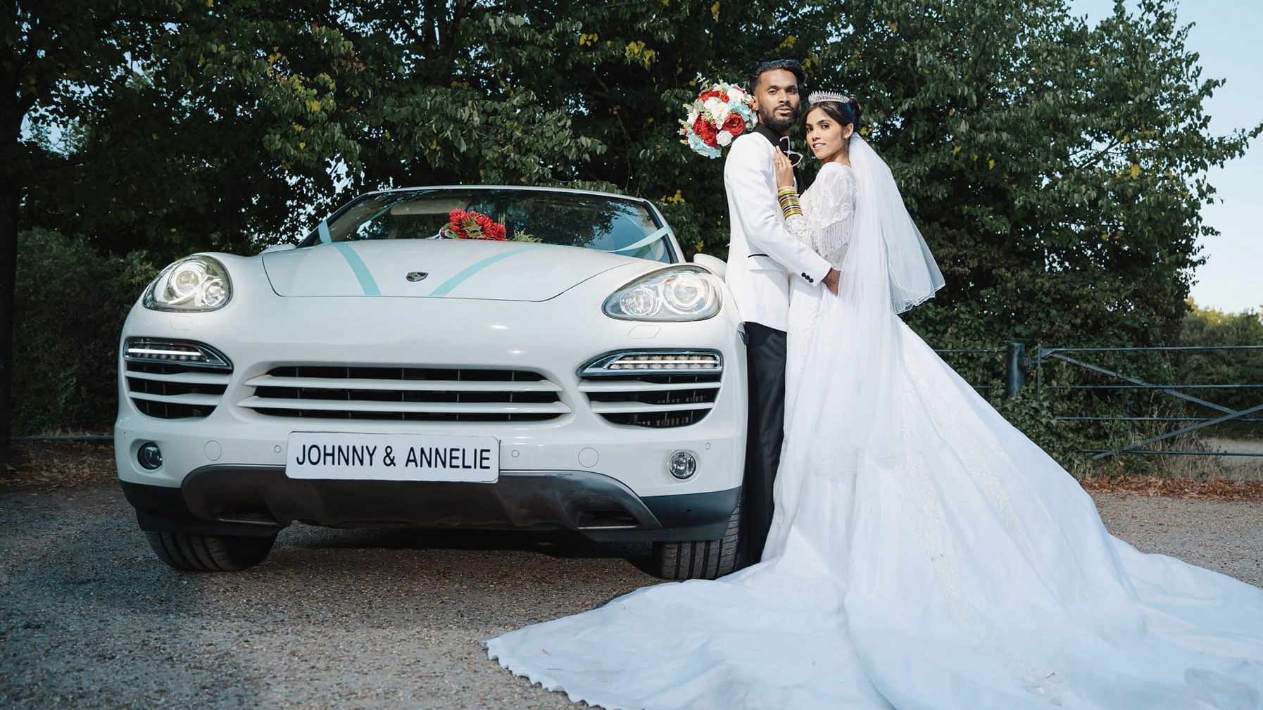 Bride and Groom standing next to White porsche Cayenne decorated with light blue ribbons. Bride is wearing a large white dress and groom a suit with black trousers and white jacket.