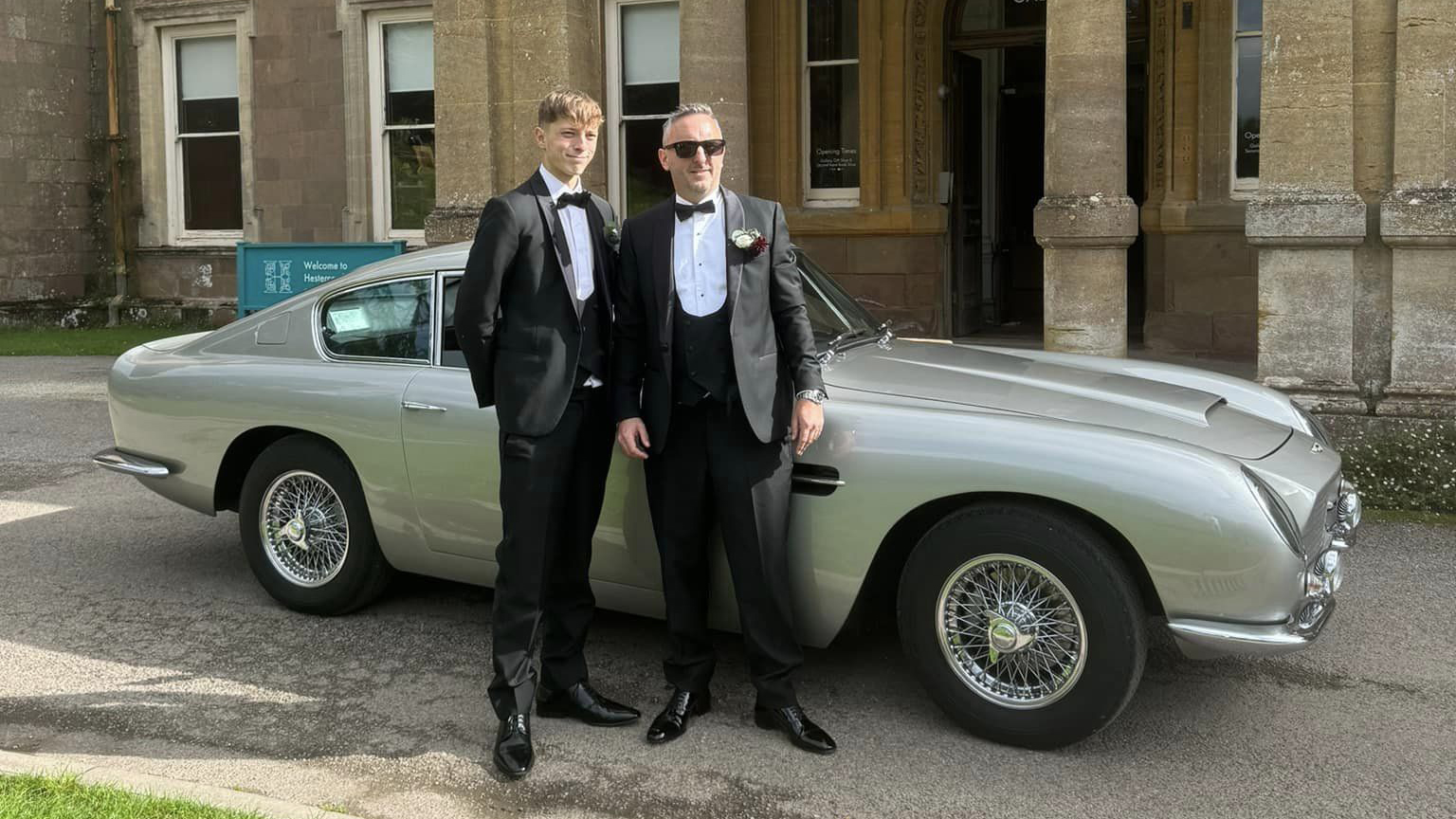 Groom and Best man wearing black wedding suits in front of Aston Martin DB6