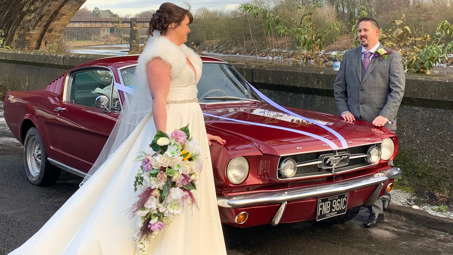 Bride wearing a white wedding dress and holding a bouquest of flowers on the right side of a classic Ford Mustang in Cherry-red with Groom on the left side of the vehicle