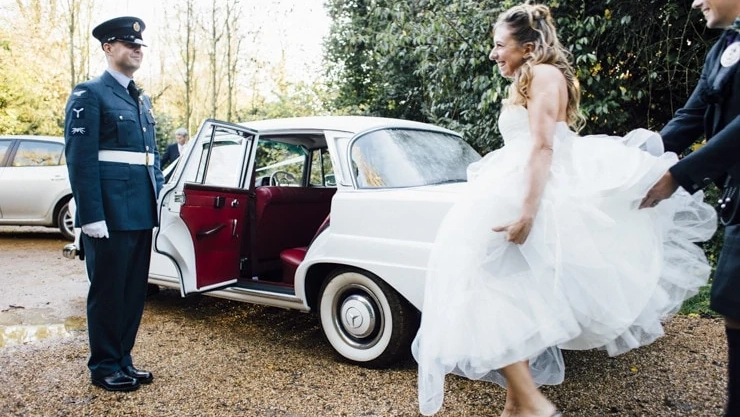 Groom in army uniform holding the door open of a classic white mercedes for its bride