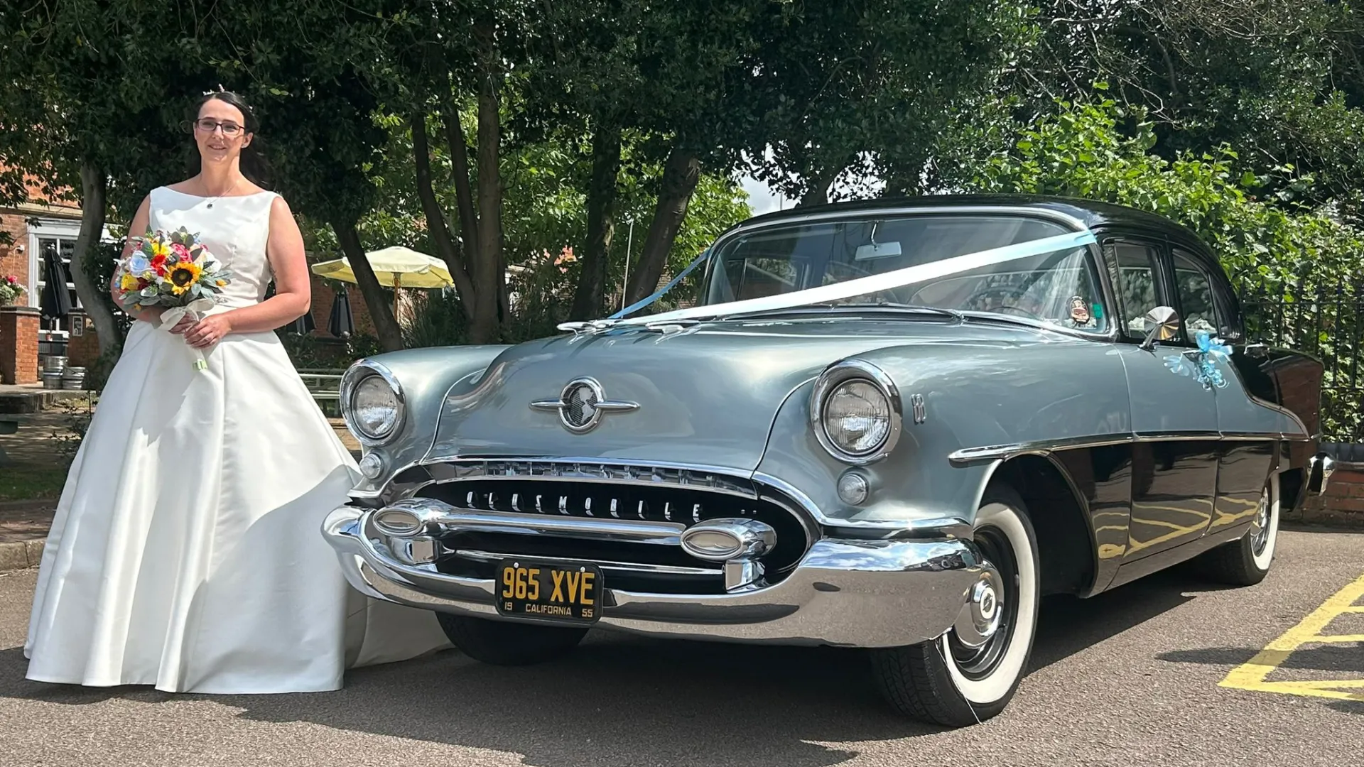 Bride wearing a white dress and holding a bouquet of flowers standing in front of a classic American Oldsmobile