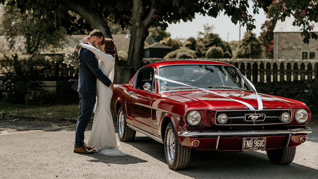 Bride and Groom holding each others next to a Classic Ford Mustang in Cherry-Red dressed with white ribbons