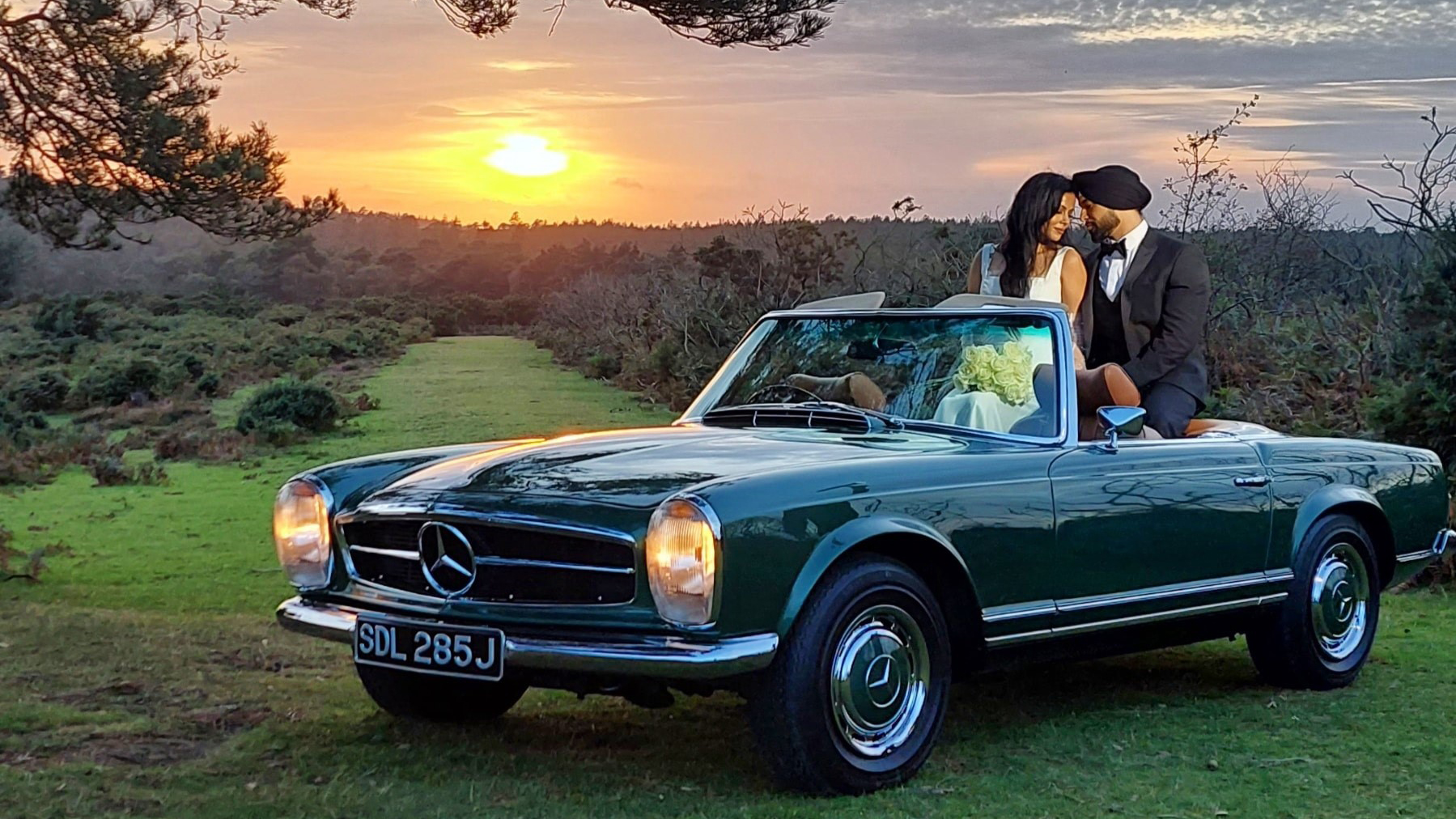 Bride and Groom inside the a classic Mercedes Convertible parked in a green garden with sunset in the background