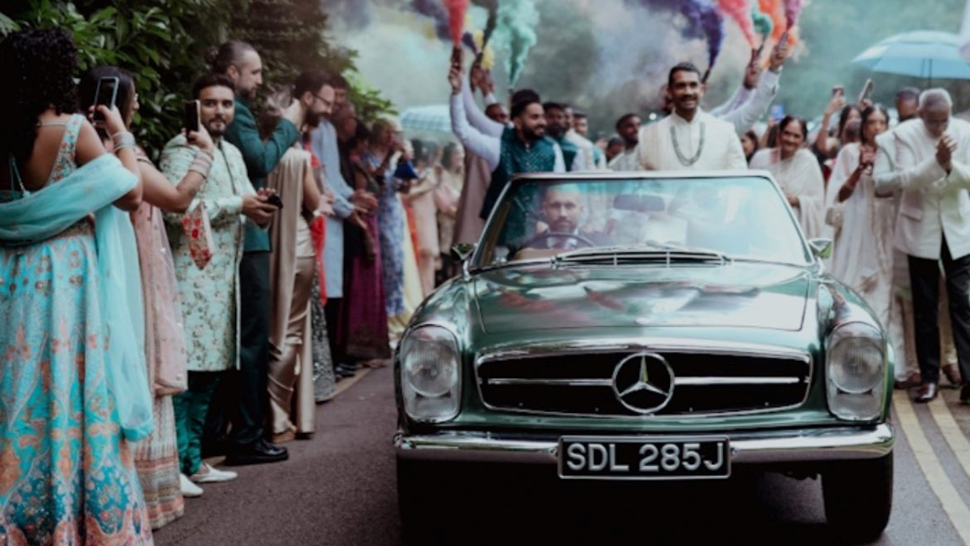 Front view of a Classic Mercedes Convertible driven by chauffeur with Asian Groom at the rear doing its wedding entrance with coloured flairs and wedding guests wearing traditional Asian outfits on both side of the vehicle