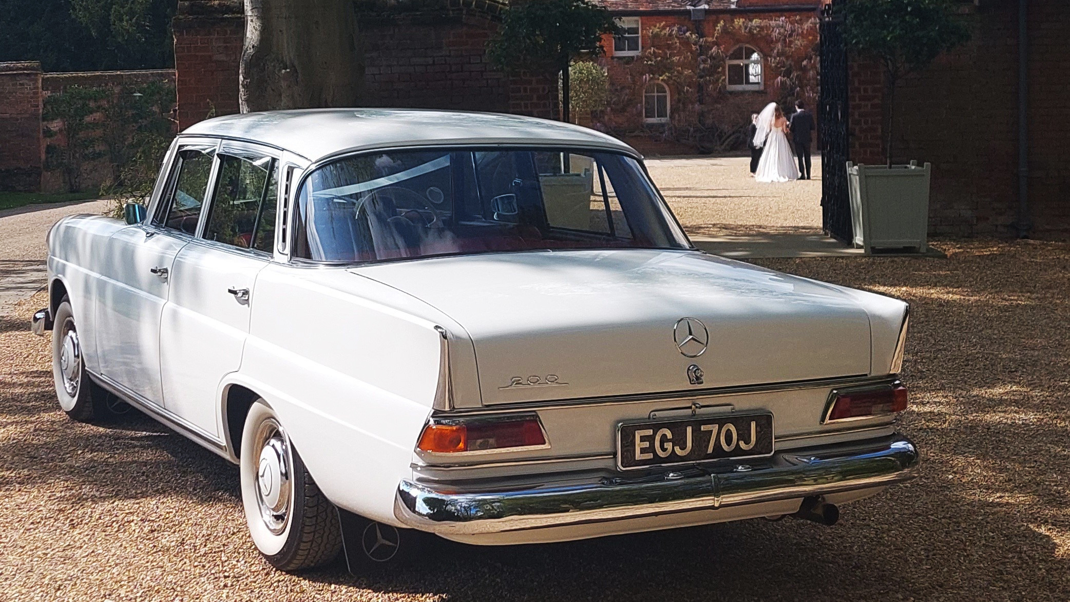 Rear view of classic white Mercedes parked in front of a wedding venue with Bride and Groom walking in the far background