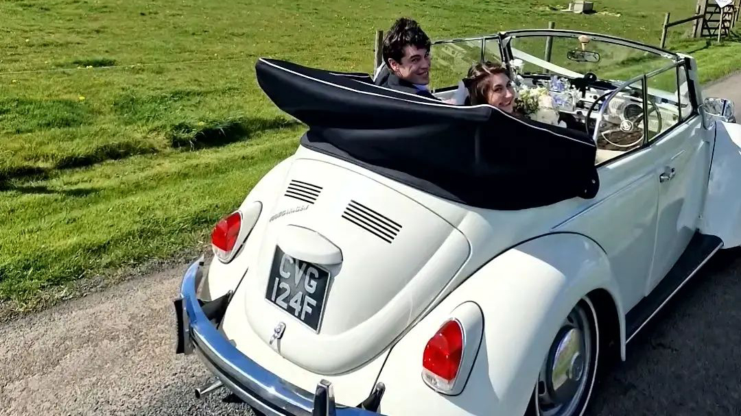 Rear view of VW Beetle with black soft top roof open. Bride and Groom seating inside the vehicle