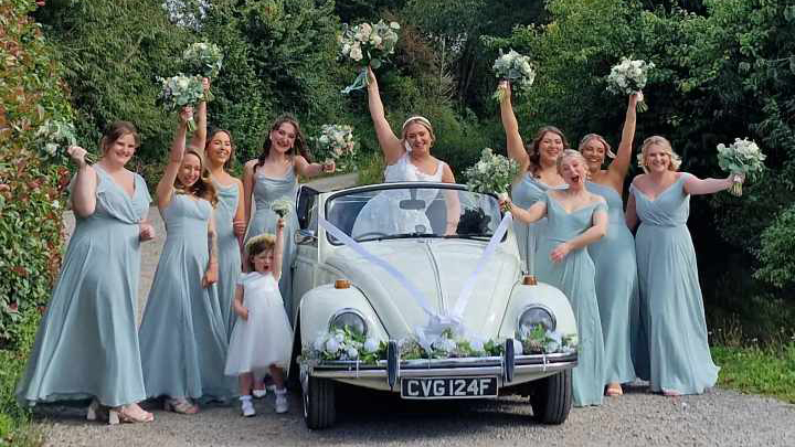 Bridesmaids wearing light blue dressed cheering around a Classic Beetle. Bride inside the vehicle standing with her White wedding gown holding a bouquet of flowers in the air.