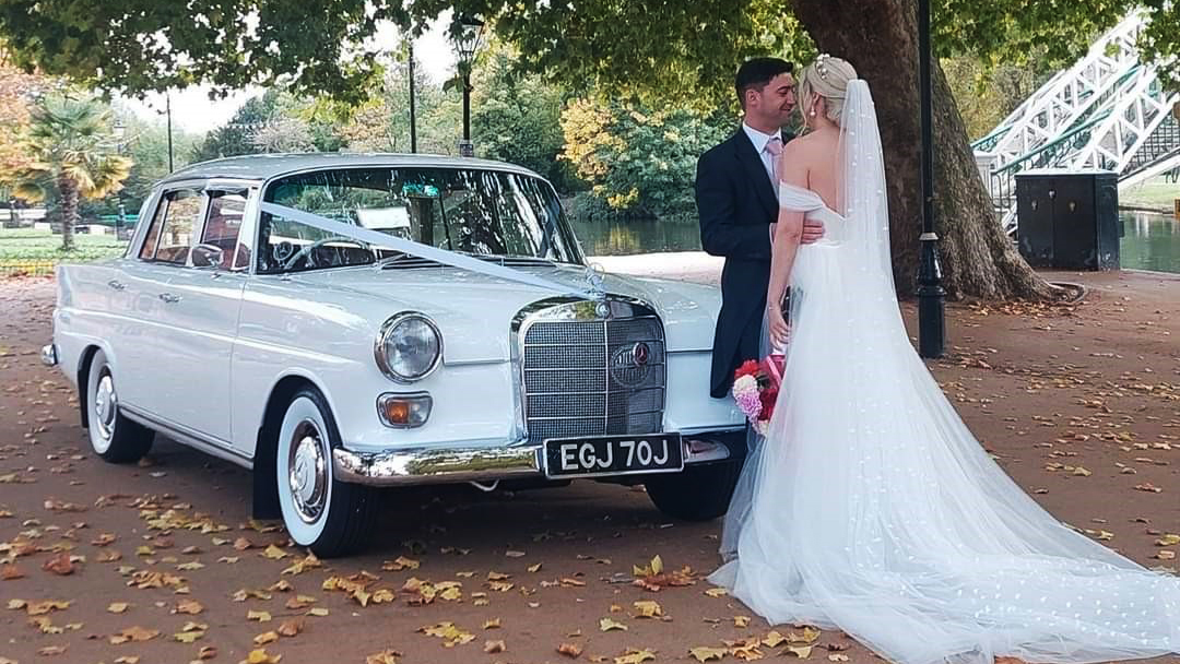 Bride and Groom holding each others in front of a White Classic Mercedes dressed with wedding ribbons