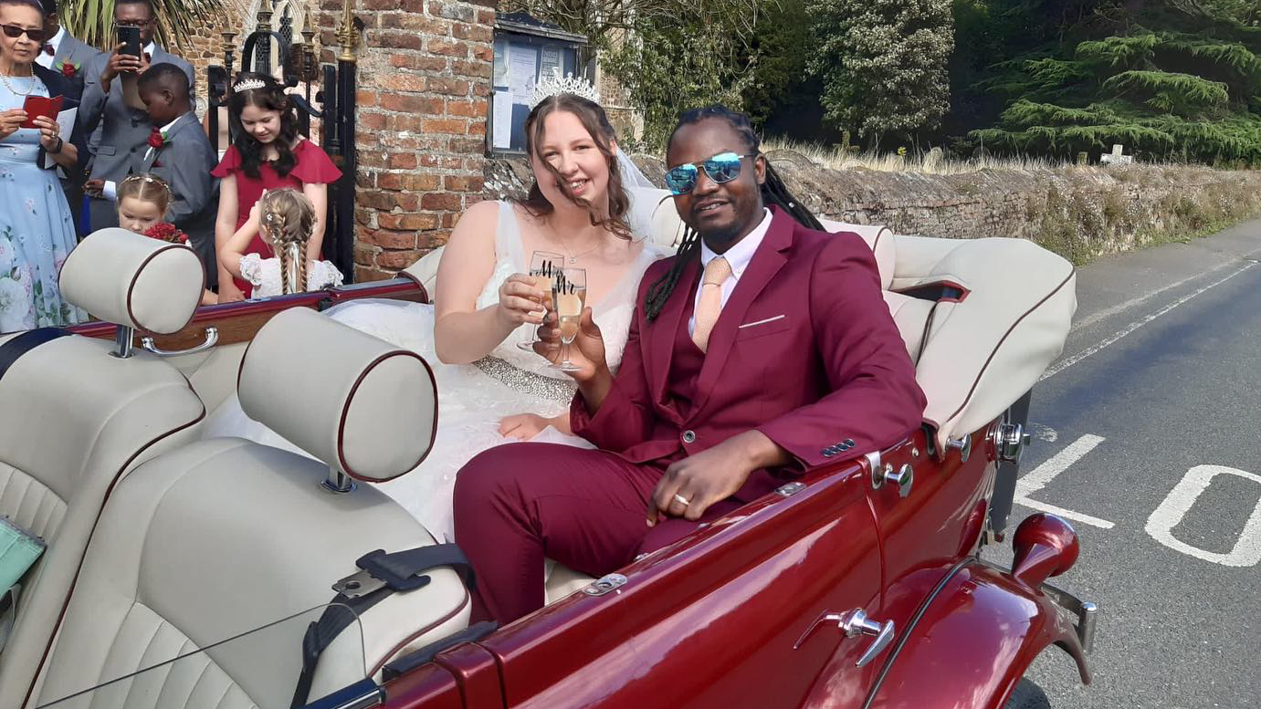 Bride and Groom seating inside a Burgundy vintage style Beauford with roof down. Wedding guests can be seen in the background.