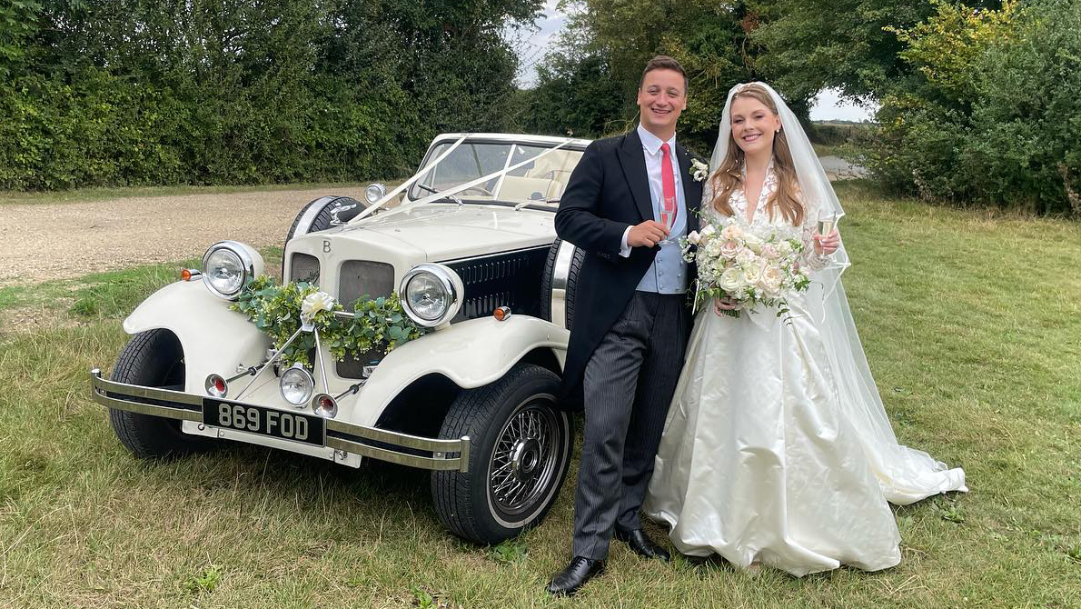 Bride holding a large bouquet of flowers in her hands with her groom standing on the left of a vintage Beauford in Black & Ivory