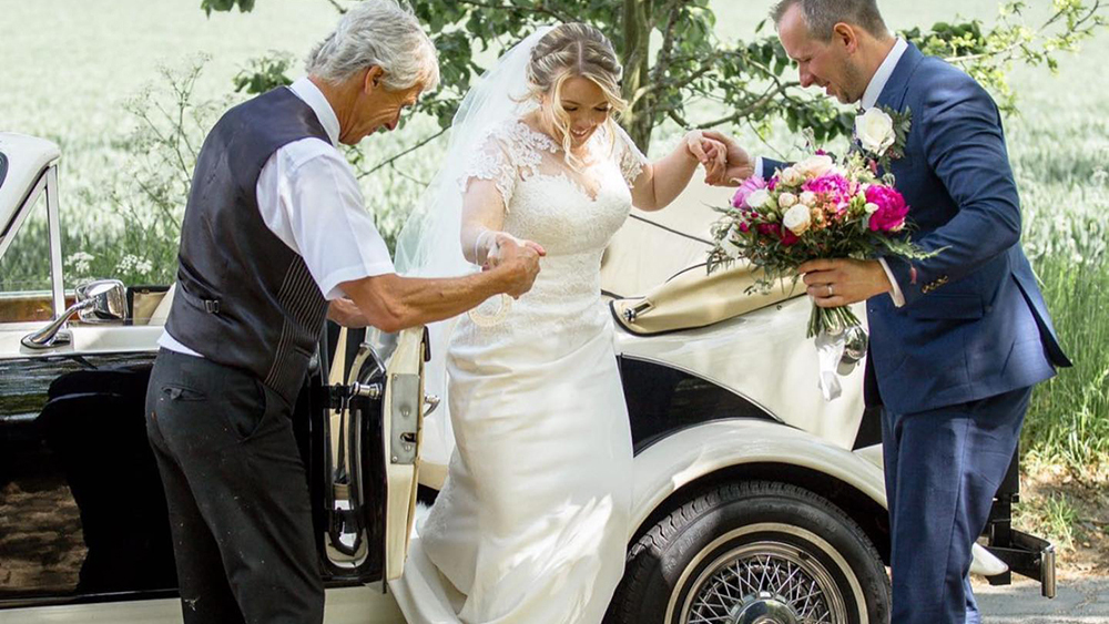 Bride exiting Beauford convertible with help from her father and her groom.