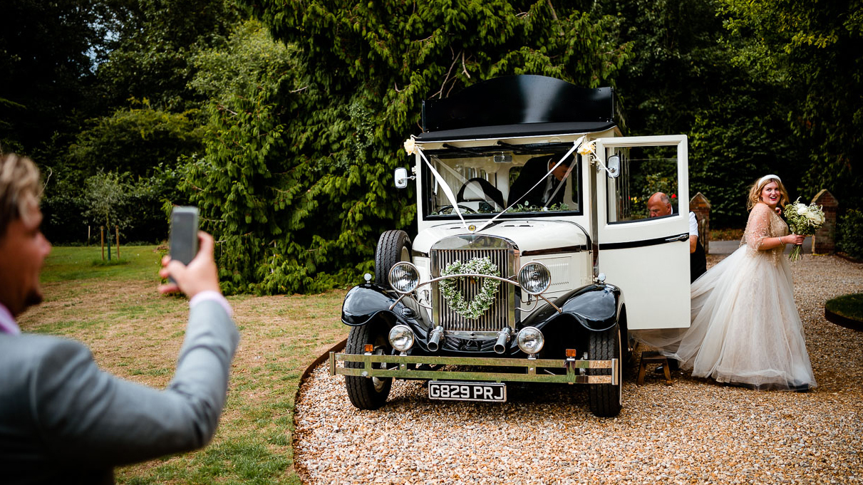 Bride wearing a White Dress exiting a Vintage style wedding bus decorated with white ribbons, wedding guests on the left taking a photo from his phone.