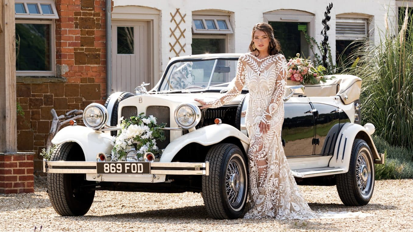 Vintage Beauford in Black & ivory with convertible roof down, flower decorattion on front chrome bumper and bride standing by the vehicle with a designer wedding dress