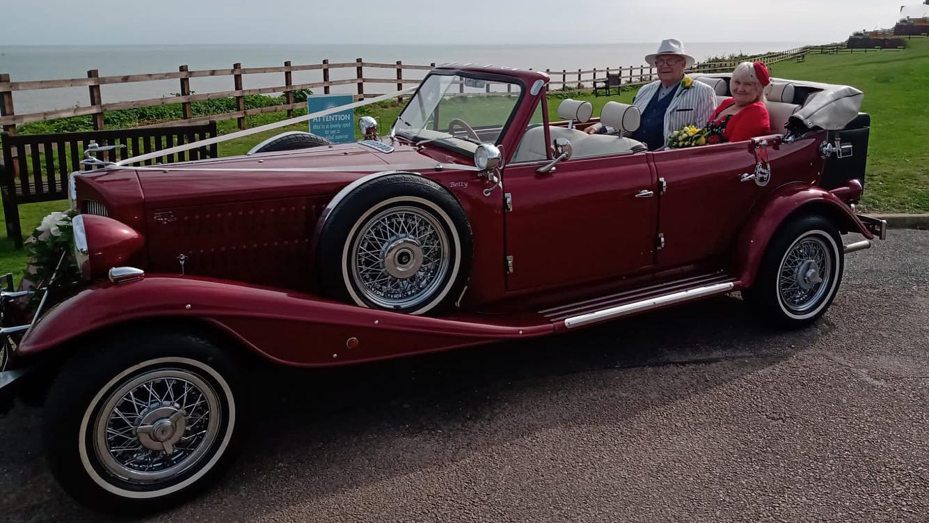 Couple seating in the rear of a Burgundy Convertible Beauford with roof down by the sea side.