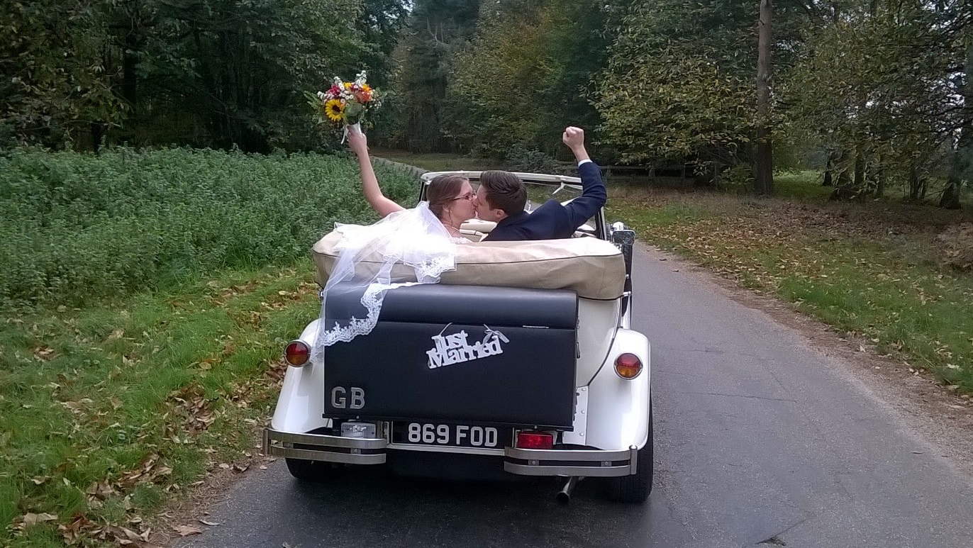 Bride and Groom seating in the back of a convertible BEauford with roof down, black pic nic trunk at the back. Couple kissing and weaving.