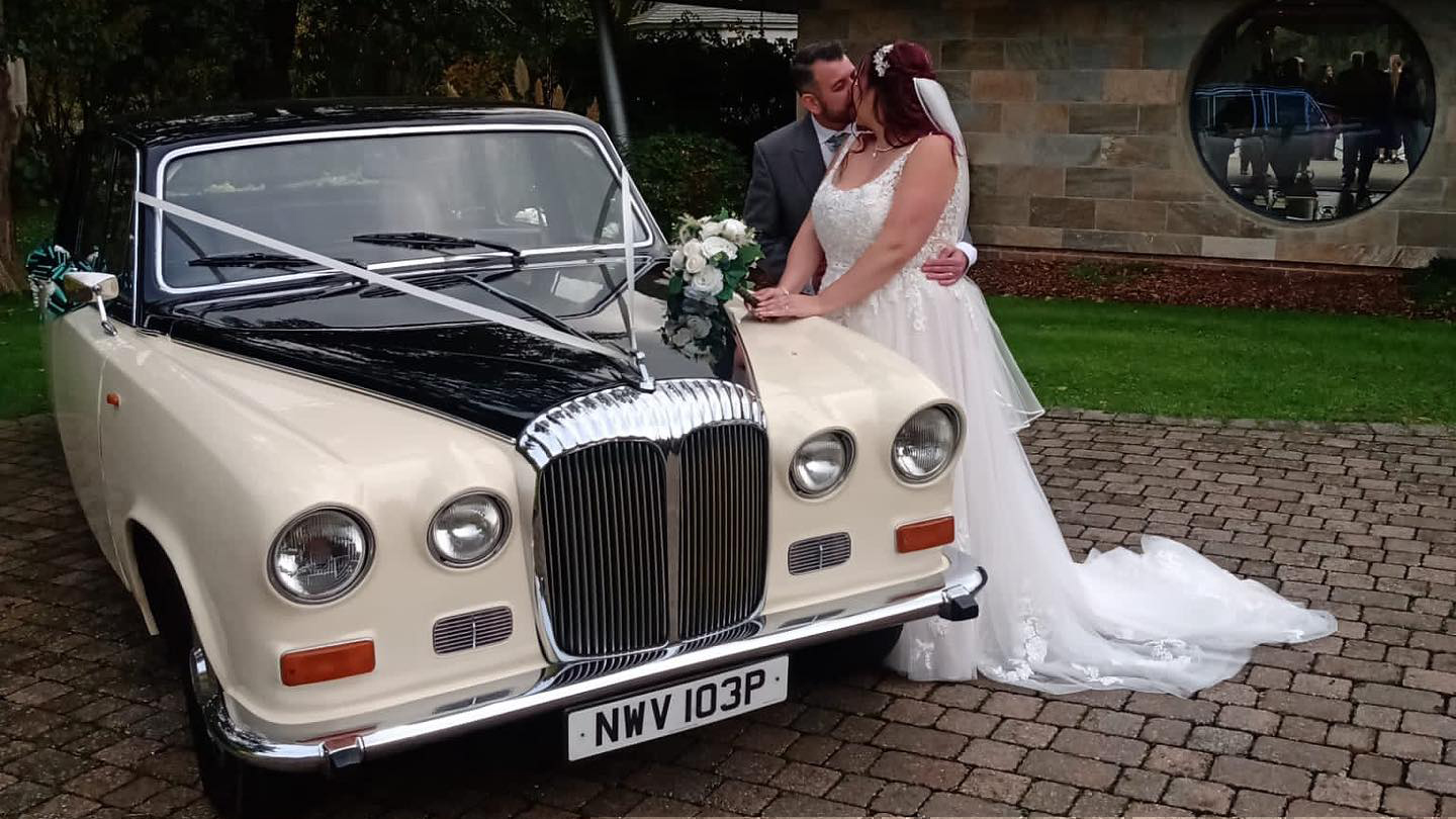 Bride and Groom kissing next to a classic Daimler limousine decorated with Ivory ribbons