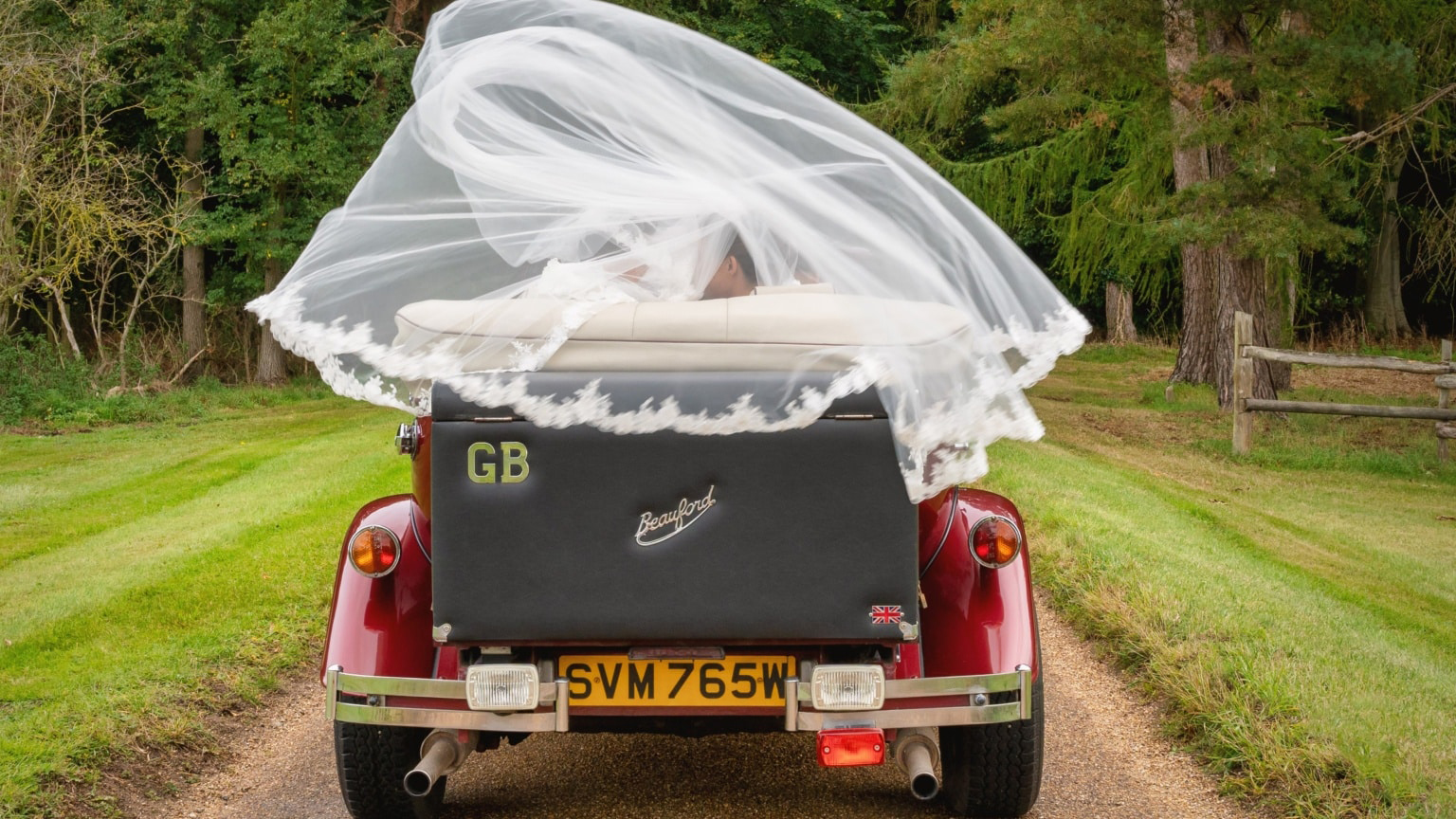 Rear view of Burgundy vintage Beauford with black picnic trunk and white wedding vale floating in the air above the vehicle.