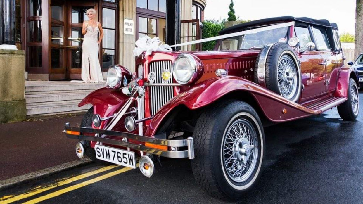 Burgundy vintage Beauford with white Ribbons and closed black soft top roof parked in front of wedding venue with Bride wearing a white dress in the background