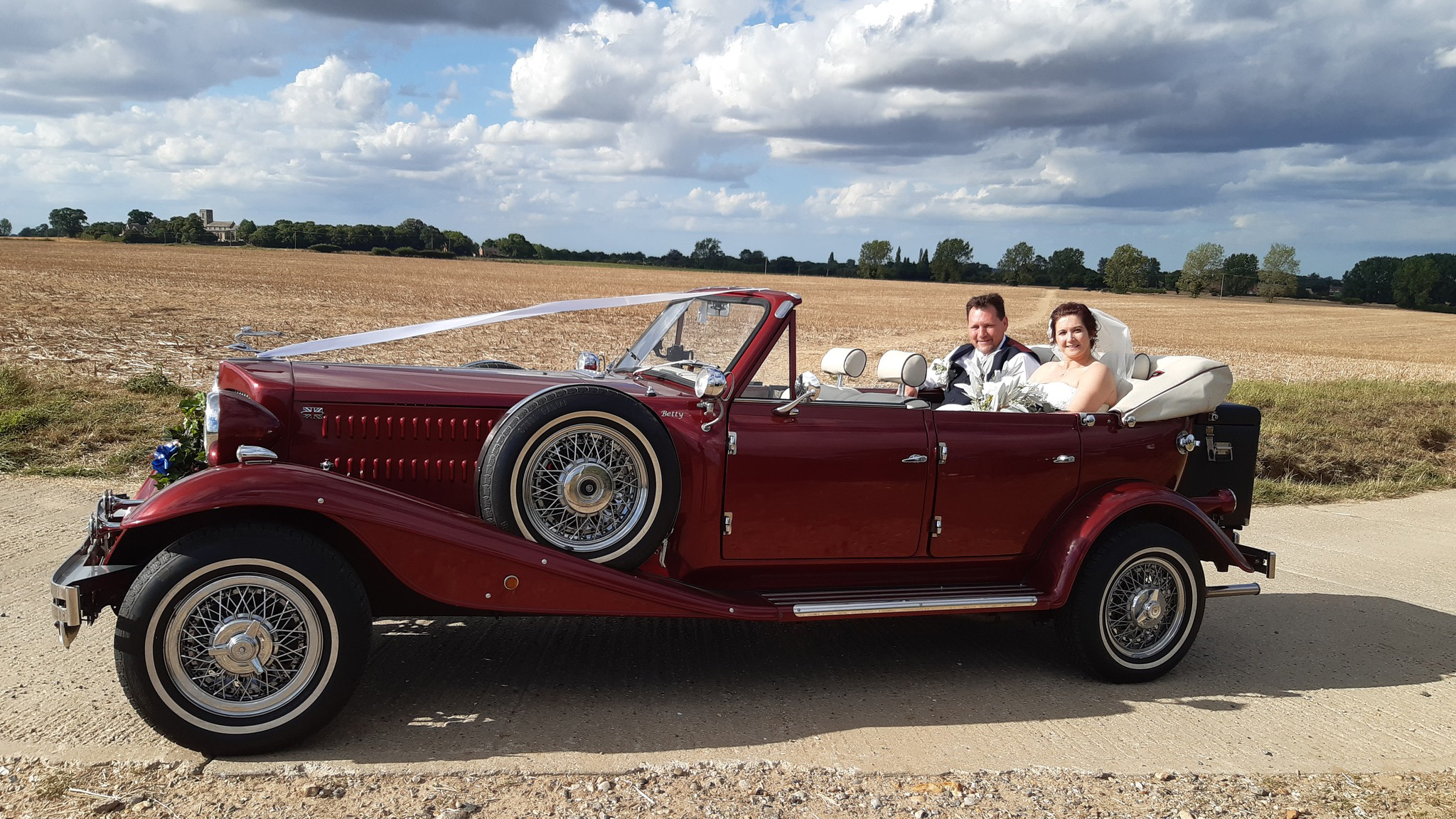 Bride and Groom seating on the rear seat of a vintage Beauford in Burgundy with roof down and decorated with White ribbons