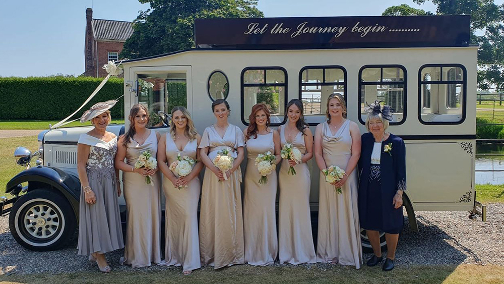 Large 8-people Bridal Party posing in front of the Vintage Asquith Wedding Bus decorated with ivoery Ribbons. Bride is wearing a qhite dress, Bridesmaids in Ivory and both mothers on with side a darker blue dresses.