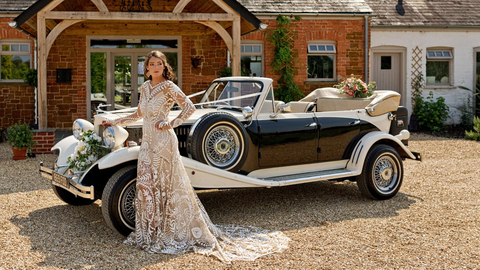 Bride wearing a designer wedding dress posing in front of vintage beauford with convertible roof down