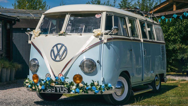 Front view of Classic VW Campervan in two tone Light blue and White decorated with Ivory ribbons and bows accross the front and some blue flowers on its front bumper.